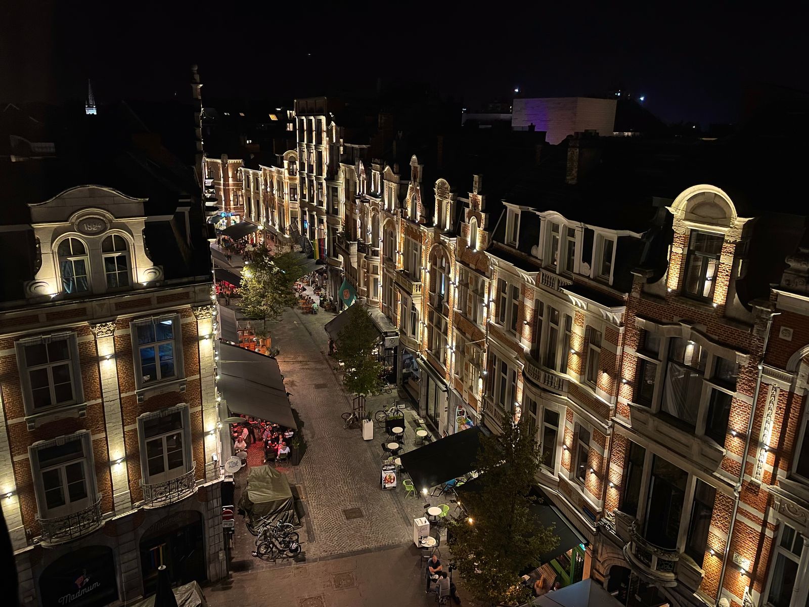 A cobblestone street at night, lit façades and café awnings, a spire in the distance