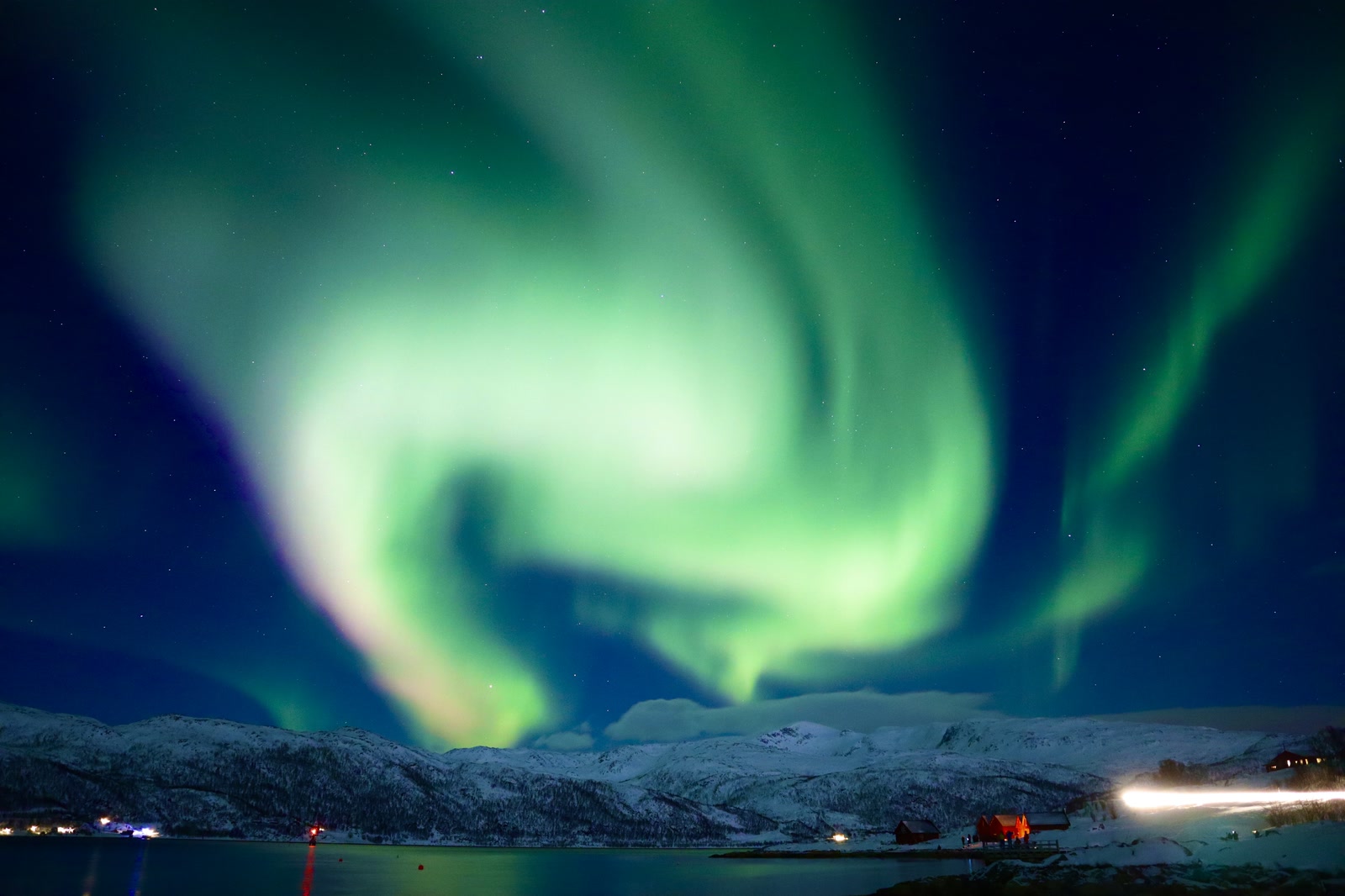 Green aurora over snowy mountains, with a lit cabin beside the water