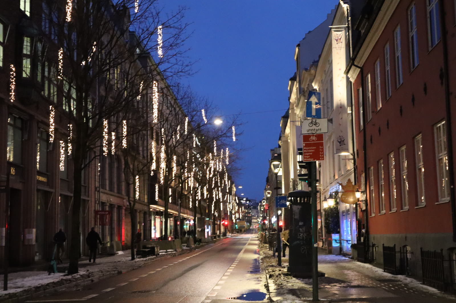 A narrow city street at blue hour, fairy lights overhead and wet pavement below
