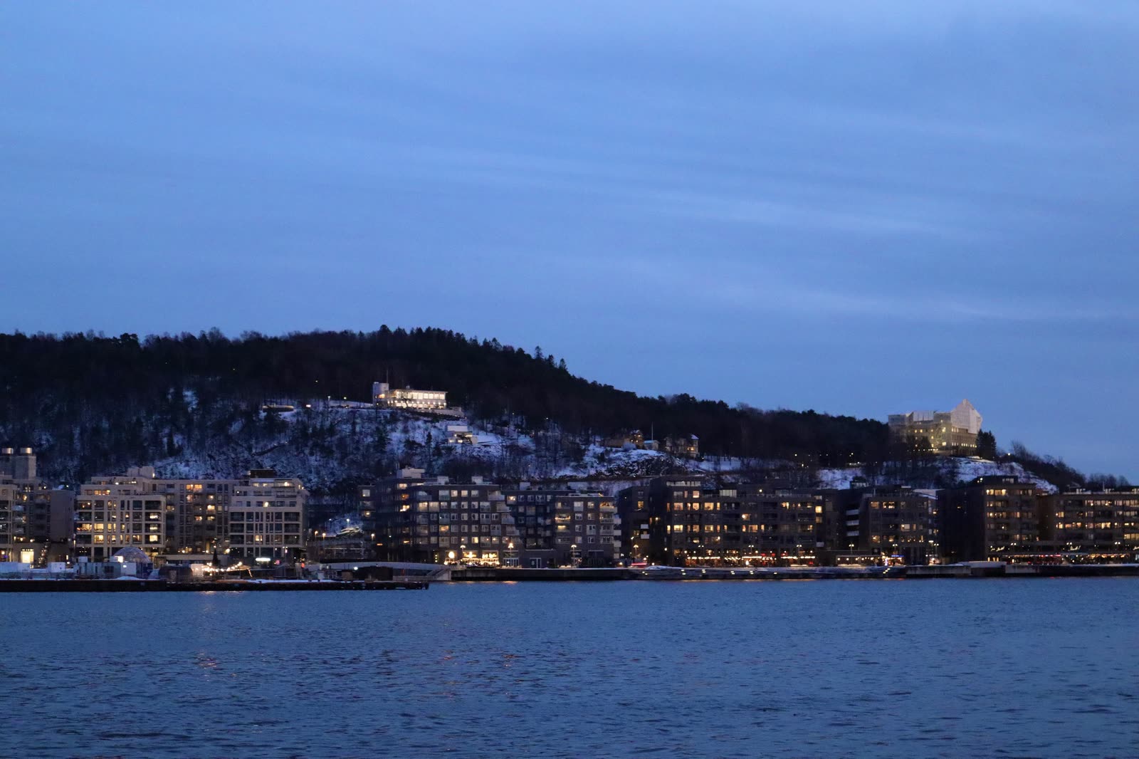 Waterfront apartments at dusk, lit windows and a snowy hillside behind, dark water in the foreground