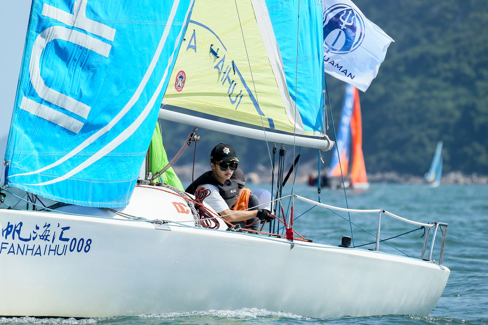 Closer on FANHAIHUI 008, blue and lime sails lettered HUI and FANHAIHUI, an Aquaman flag, and a sailor in a black vest