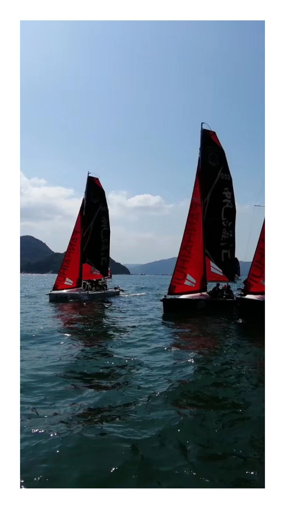 Dinghies with red-and-black sails, hull 002 visible, on blue-green water with hills behind