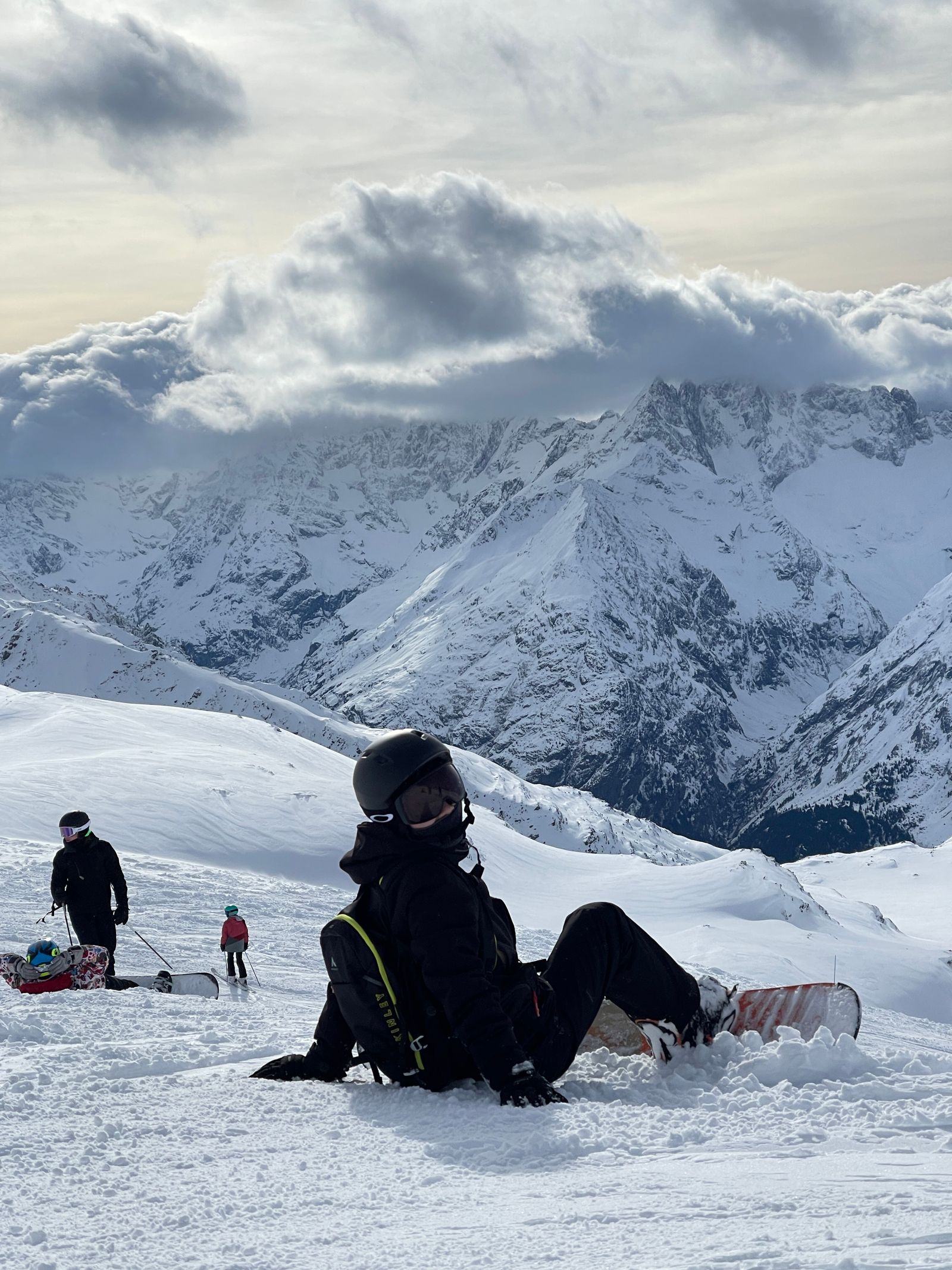 A snowboarder sitting in the snow, looking out at a rugged range under the clouds