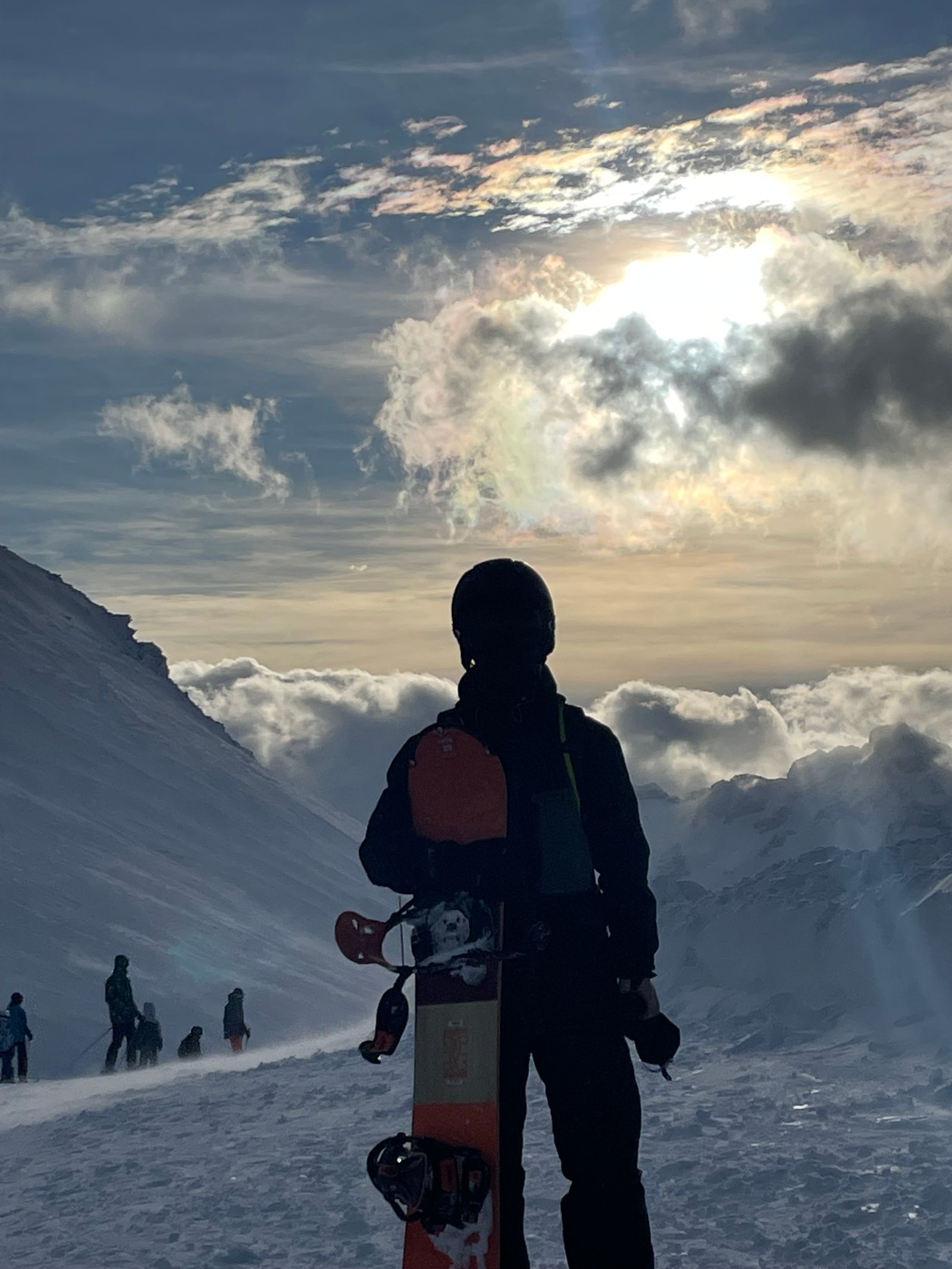 A snowboarder seen from behind, board in hand, the sun flaring through clouds above a sea of clouds