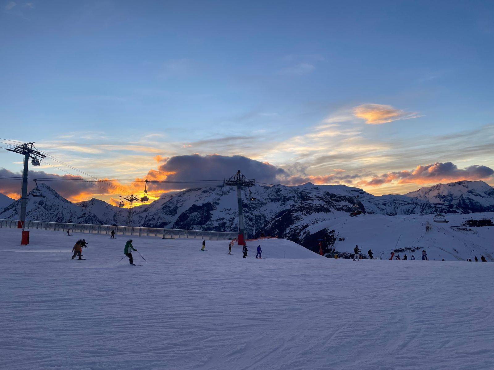 A groomed piste and chairlift towers, orange sunset behind the peaks, skiers on the slope