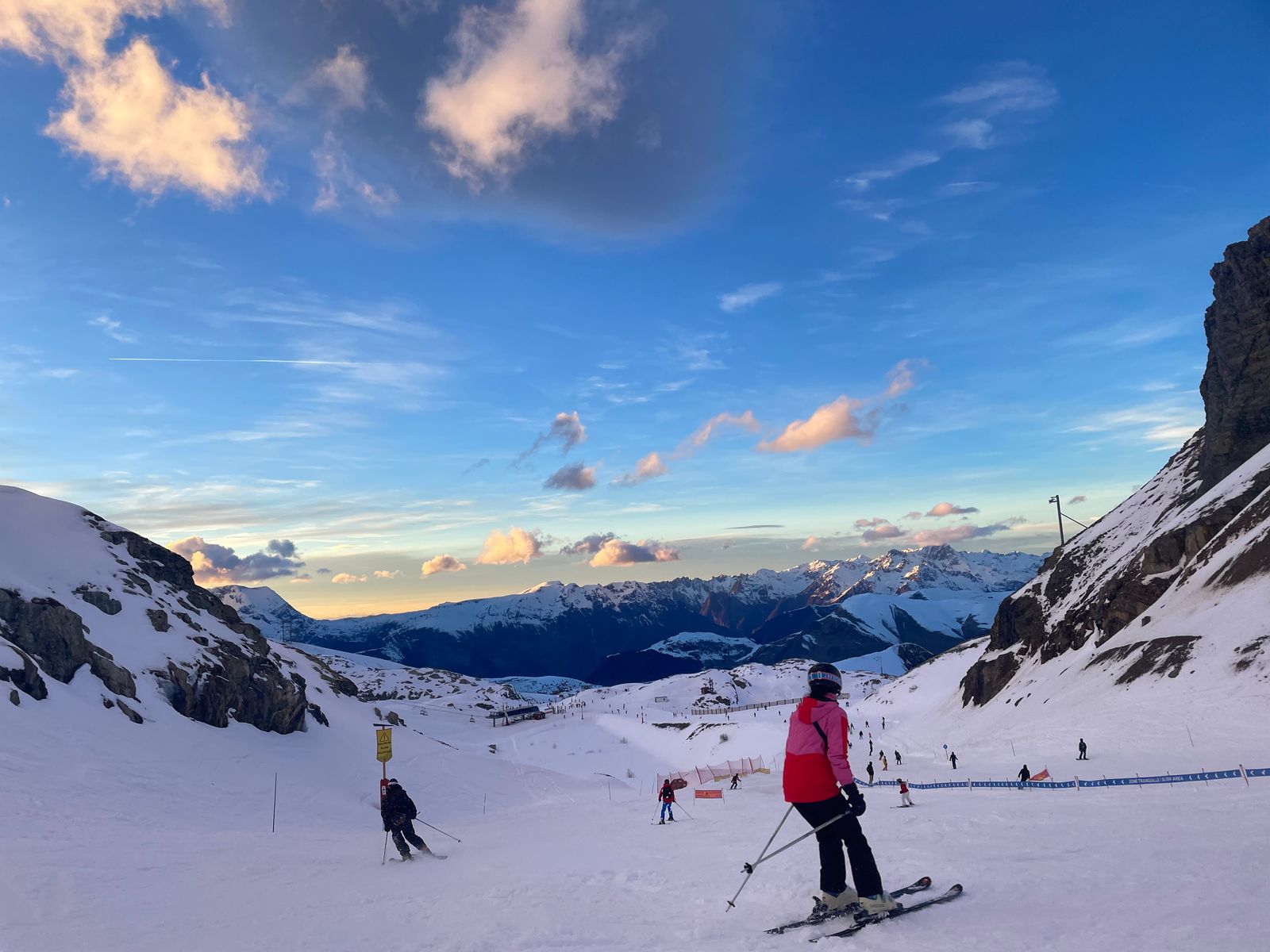 A skier in a pink jacket at the top of a valley run, golden light on the snow, peaks under a blue sky